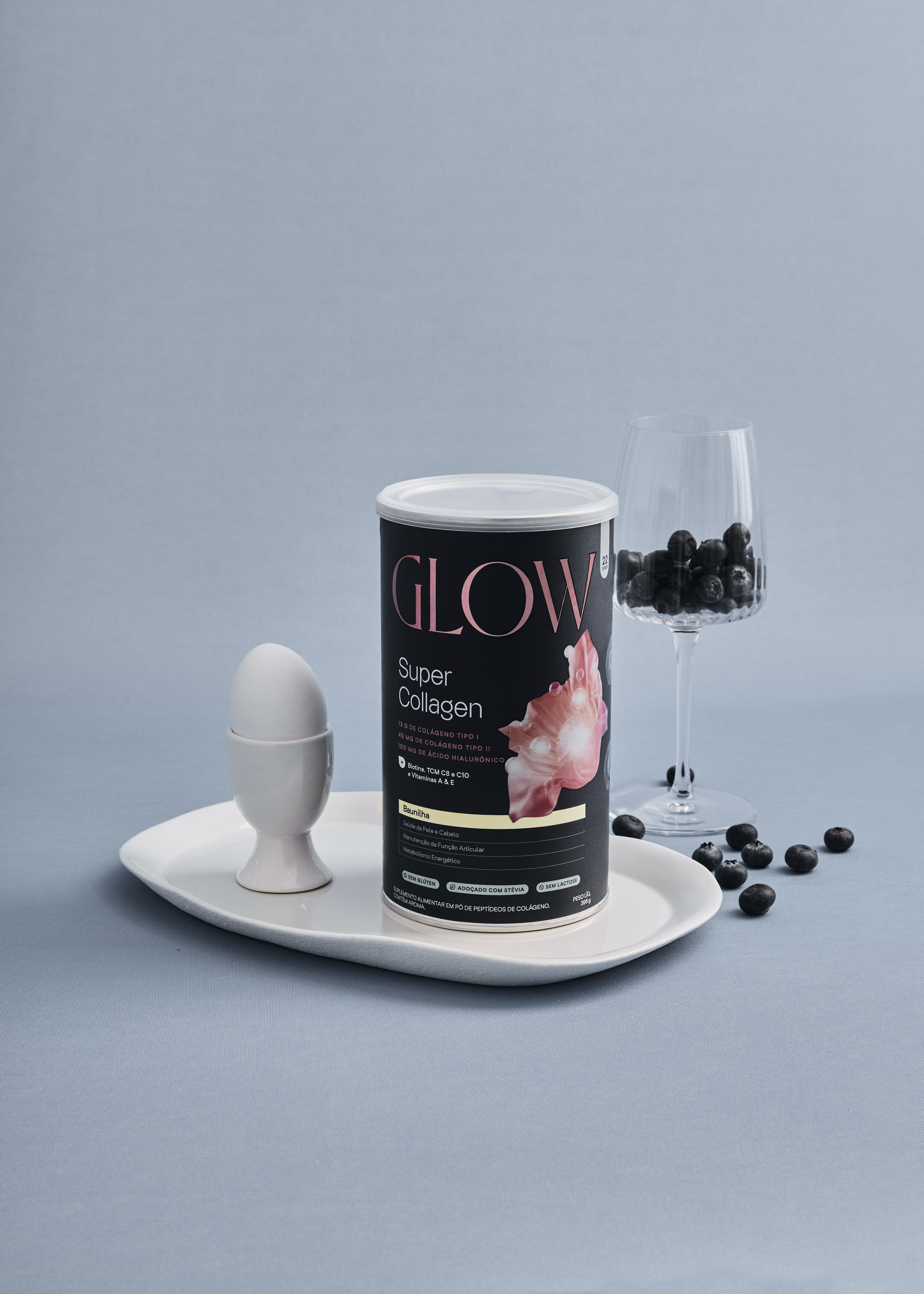 Pote do suplemento Glow Super Collagen sabor Baunilha sobre bandeja branca, acompanhado de um ovo em casqueiro branco, uma taça de cristal com blueberries e frutas espalhadas ao redor, sobre fundo azul acinzentado